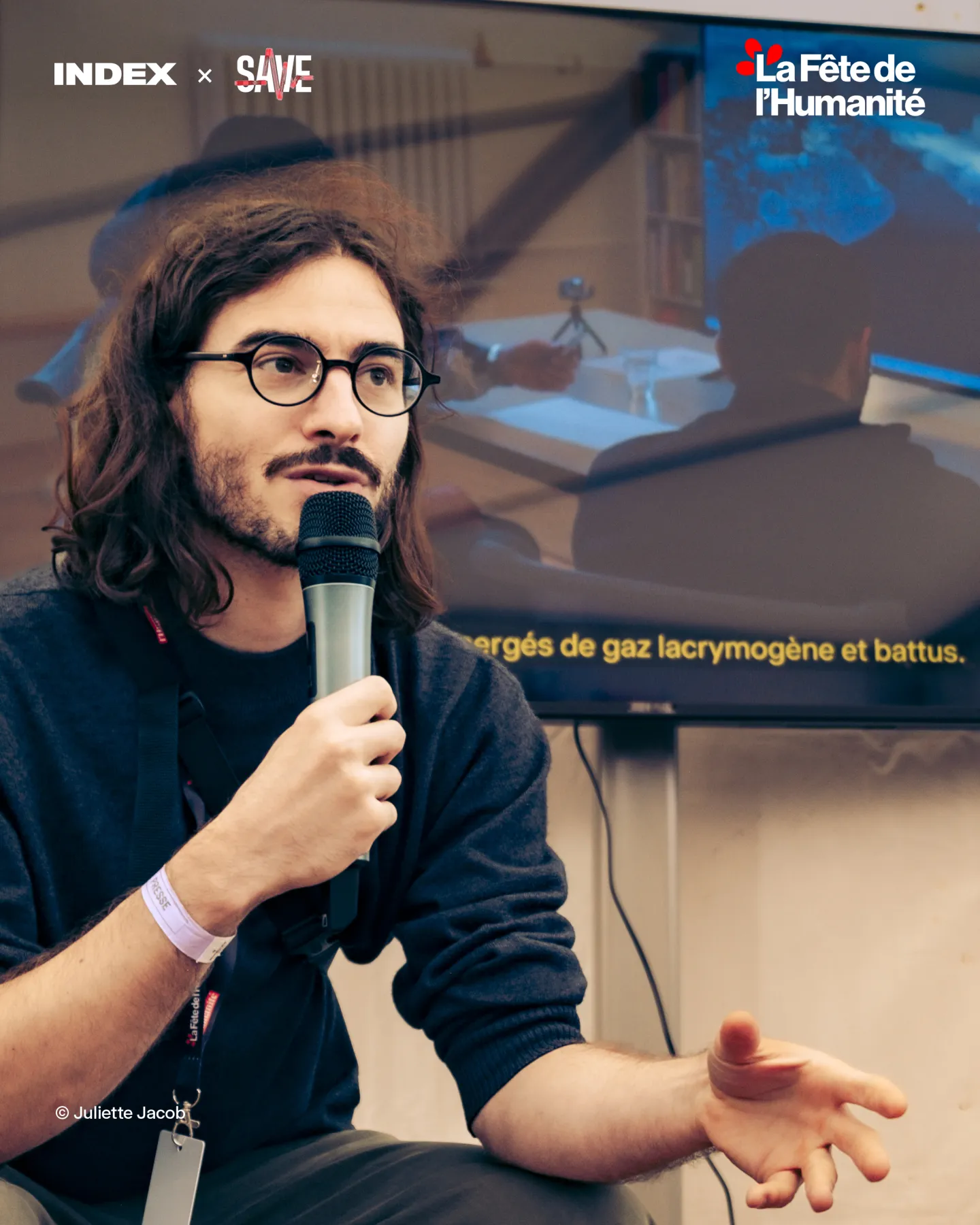 An Index speaker at the microphone in front of a screen showing a subtitled testimony