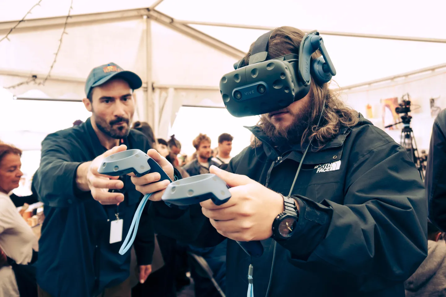A visitor wearing a virtual reality headset, controllers in hand, guided by an Index member under a marquee