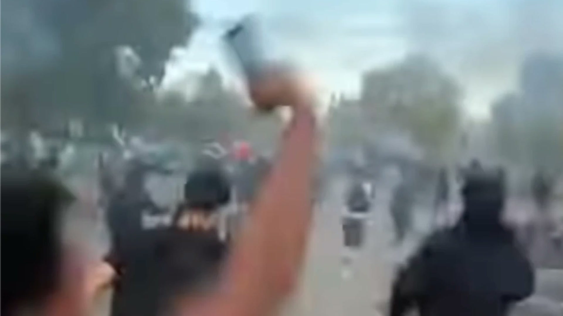 Blurred image: students running down a smoky street, a raised arm holding a phone in the foreground