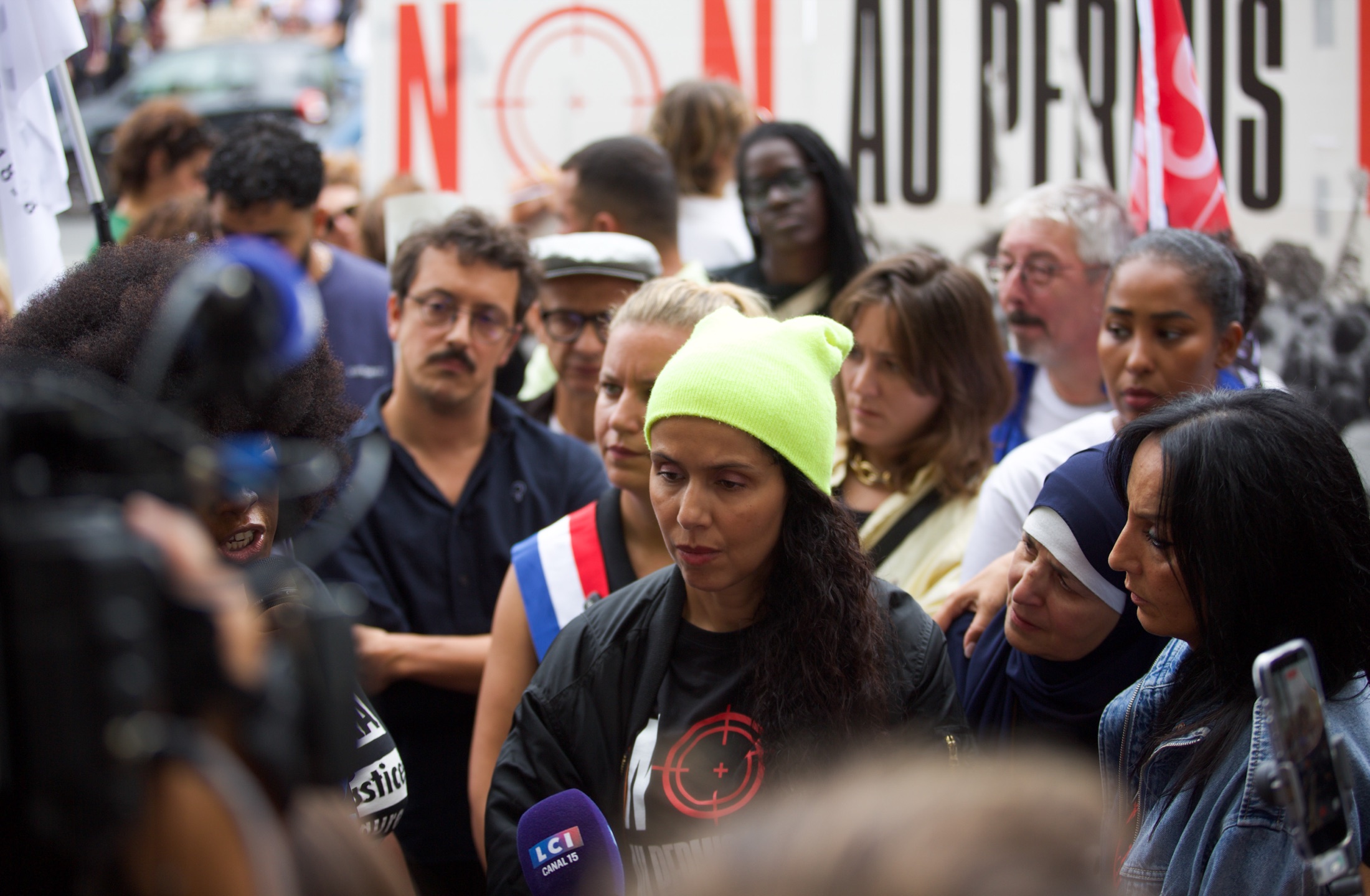 Une femme s’exprime devant une caméra de télévision, entourée de manifestant·es, devant une banderole « Non au permis… »