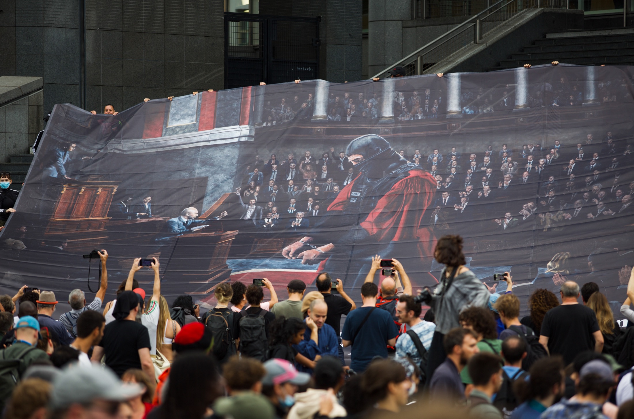 Une fresque géante déployée sur des marches représente un hémicycle parlementaire où siège un policier casqué en robe rouge ; la foule la photographie
