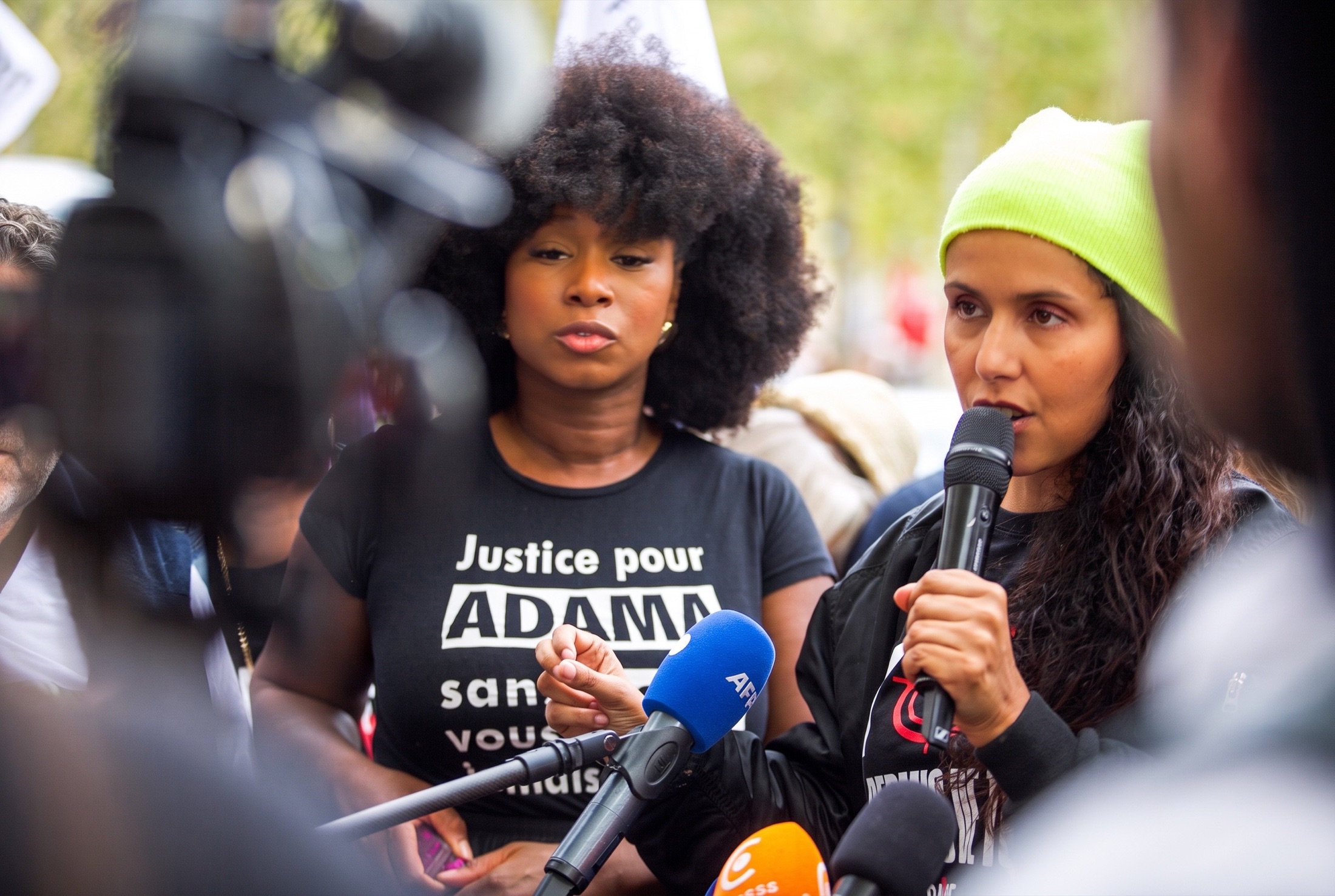 Deux femmes s’expriment devant un bouquet de micros de presse lors d’une manifestation ; l’une porte un t-shirt « Justice pour Adama »