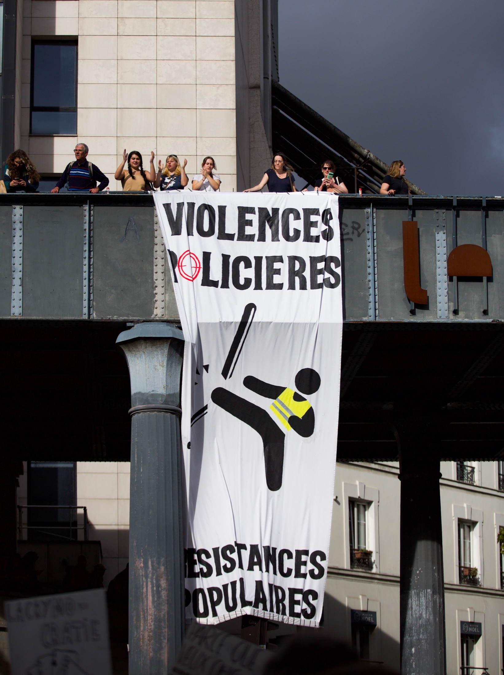 Une banderole verticale « Violences policières — Résistances populaires » déployée depuis un pont métallique